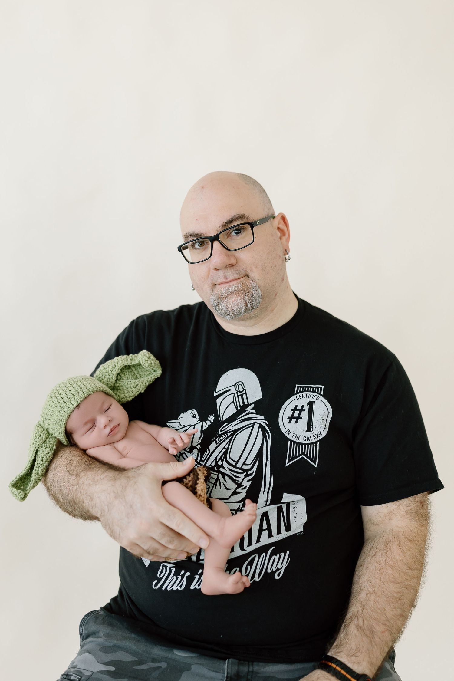 A person wearing a black graphic t-shirt holds a sleeping newborn wearing a knitted green hat against a light background.