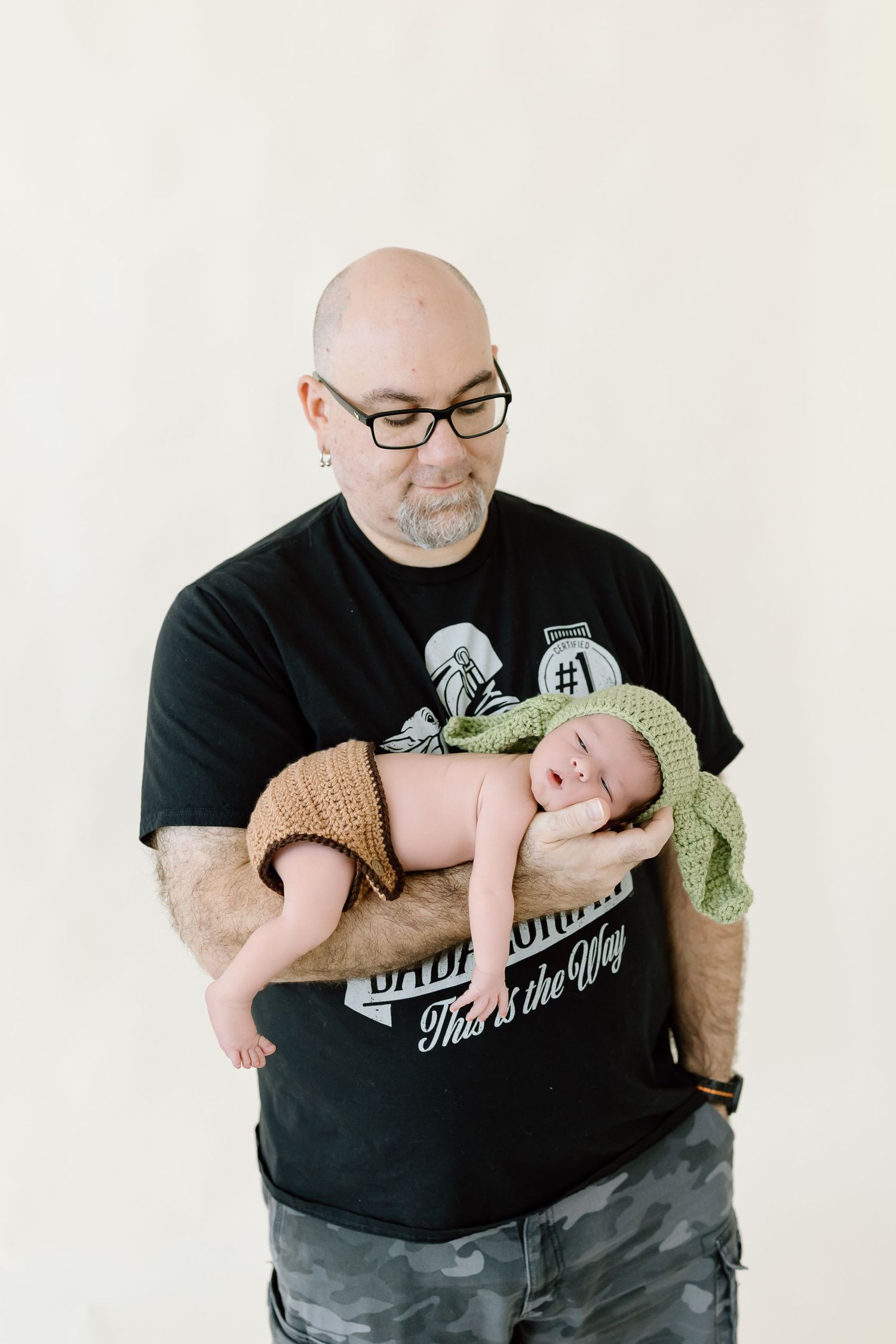 A person in a black t-shirt holds and gazes lovingly at a sleeping newborn baby wrapped in a green blanket.