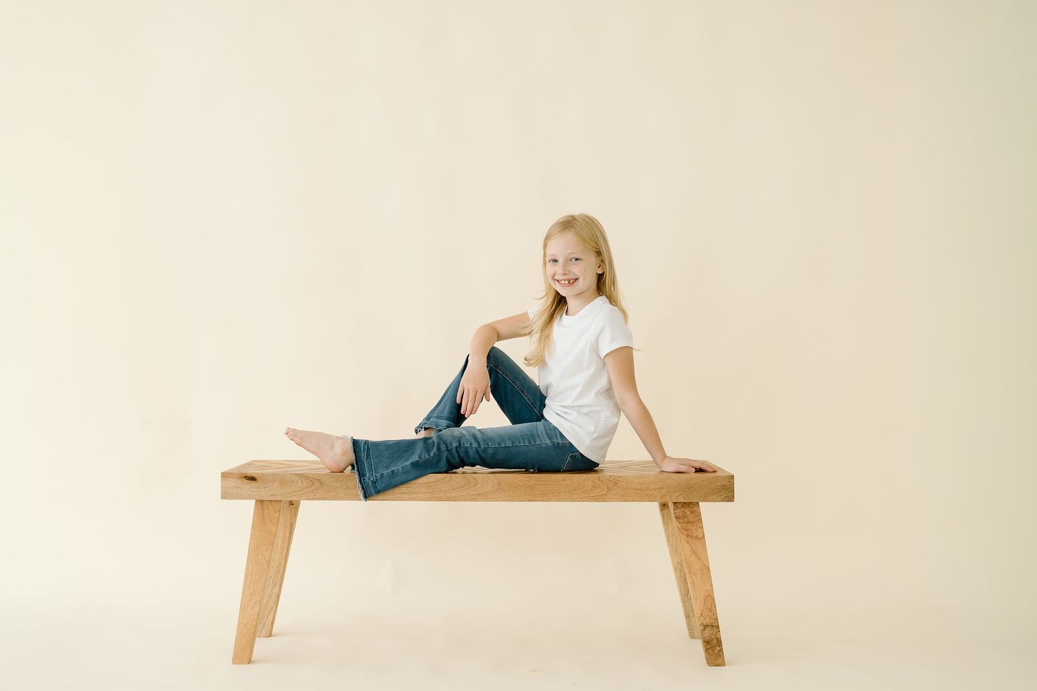 A person in white clothing lies on a wooden bench in a minimalist setting against a beige background.