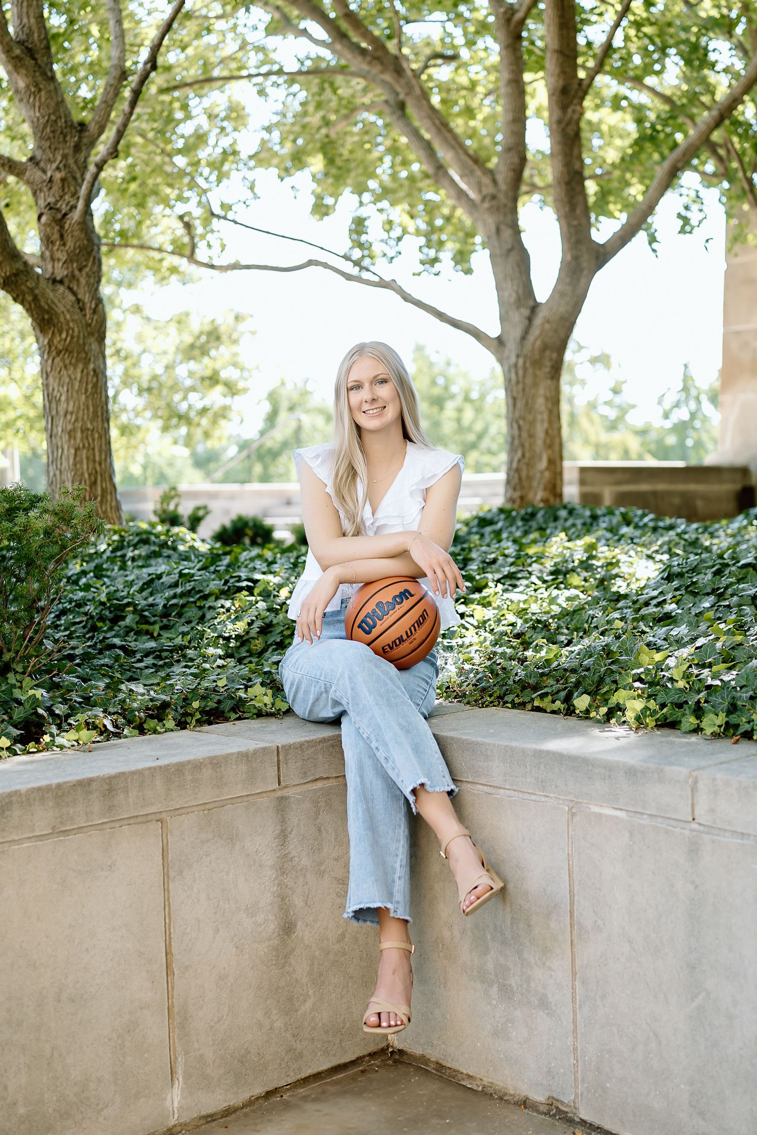 A person in light clothing sits on a concrete wall surrounded by lush green trees and foliage in a serene outdoor setting.