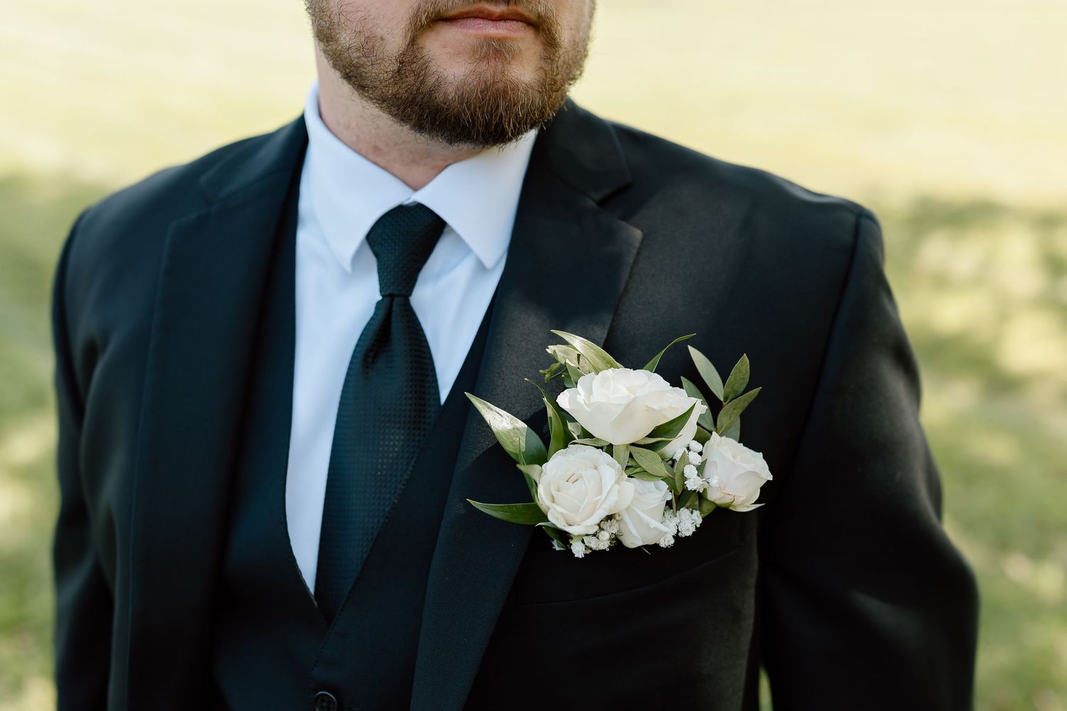 Close-up view of a black suit lapel with white rose and greenery boutonniere at a formal wedding ceremony.