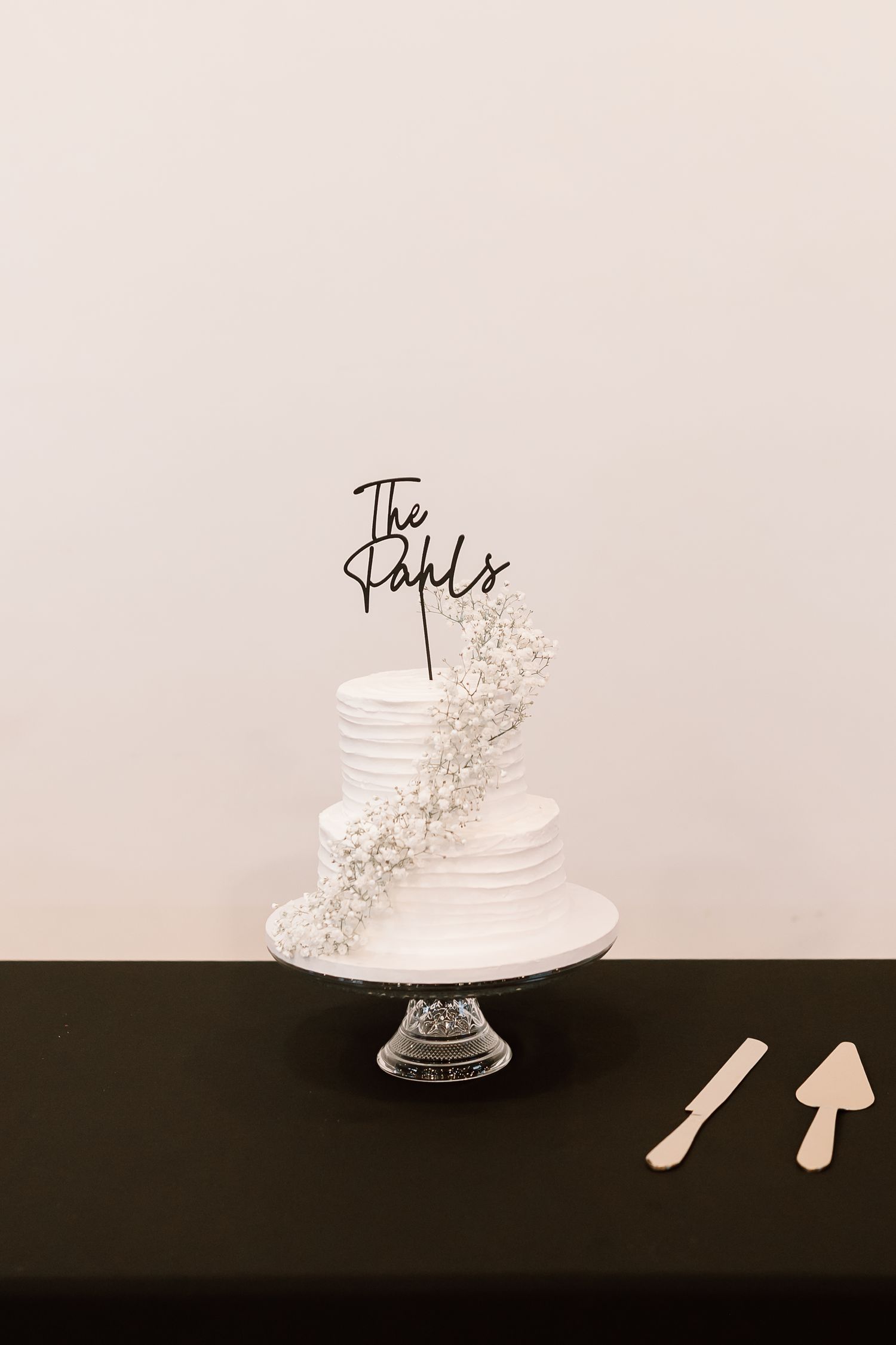 Elegant white wedding cake with delicate floral decorations displayed on a clear glass pedestal against a dark background.