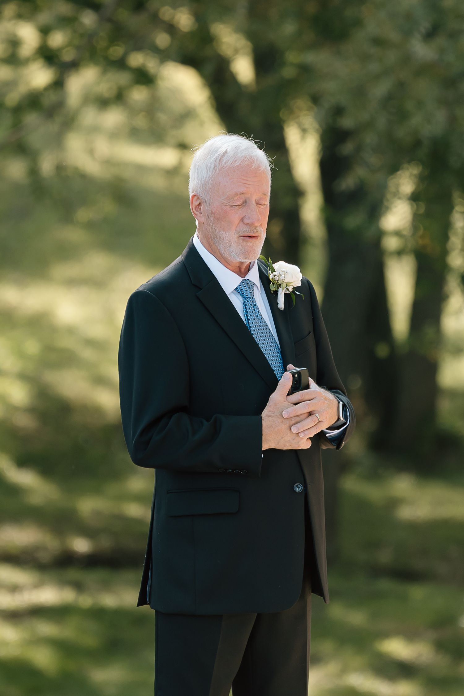 Man in black suit and tie checks mobile phone while standing outdoors in a sunlit wooded area.
