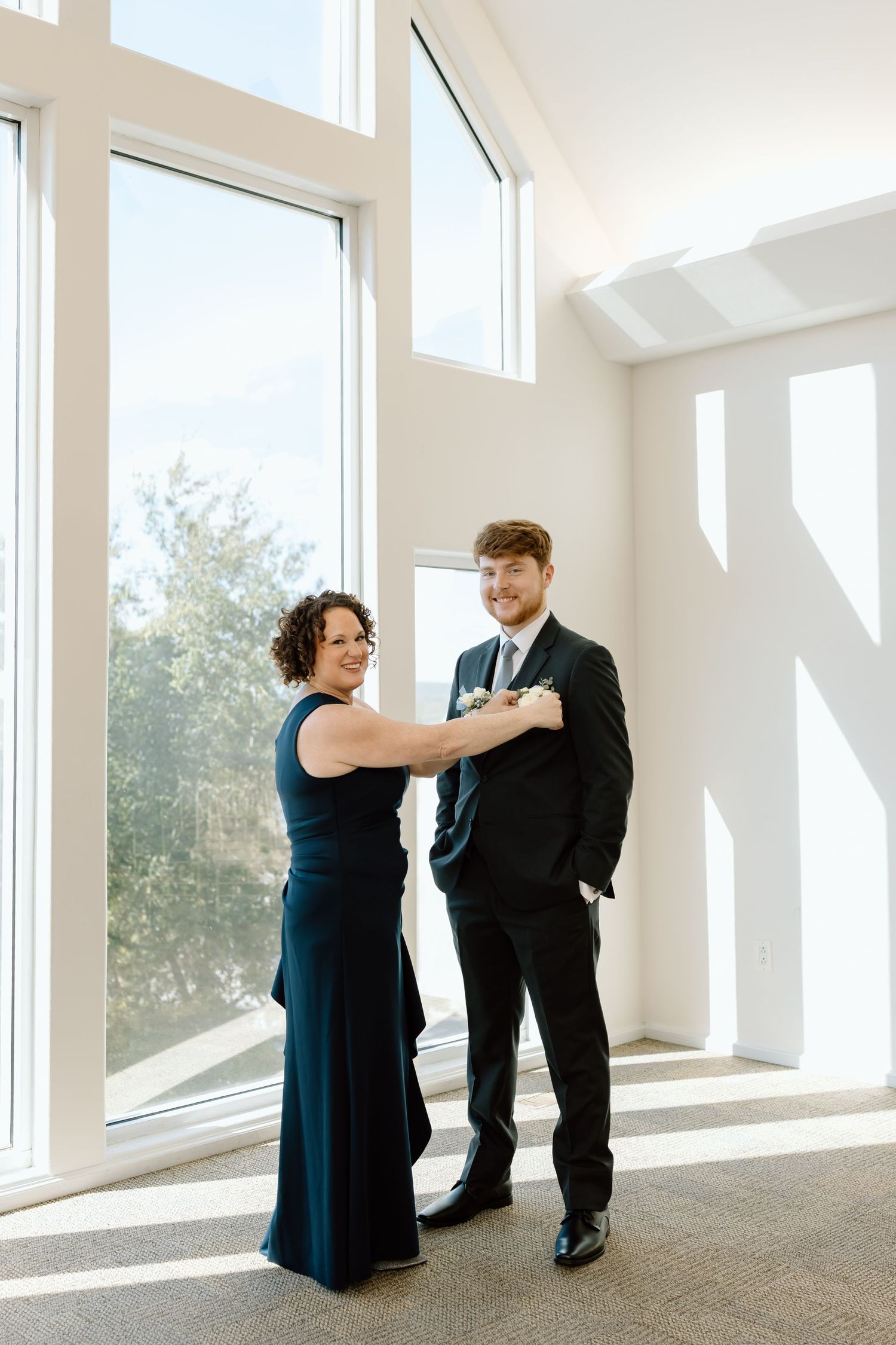 Two people in formal attire share a tender moment in a bright, modern room with floor-to-ceiling windows.