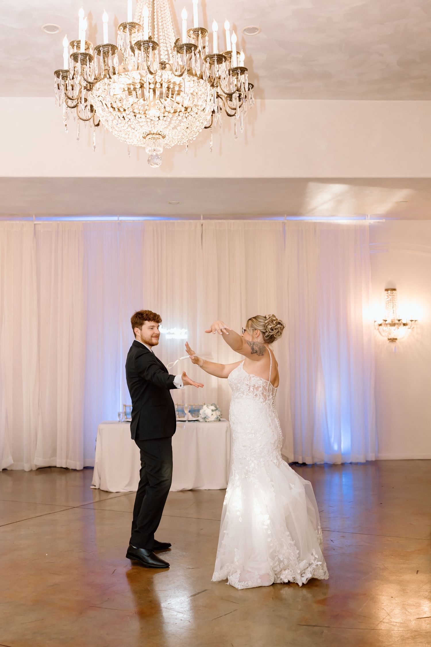 Couple sharing their first dance in a luxurious white-draped ballroom with sparkling chandeliers overhead.