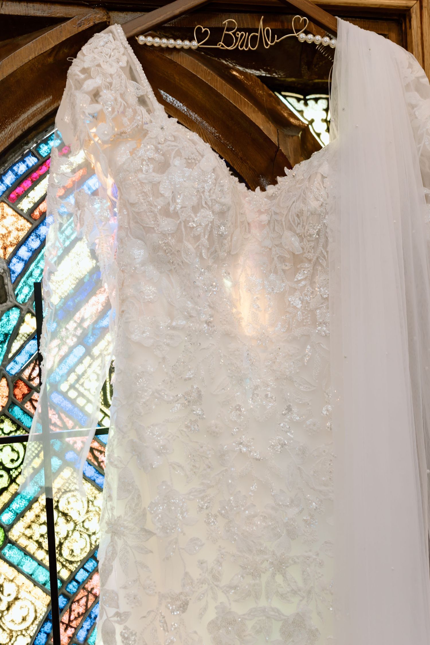 White wedding dresses with intricate lace details hanging on display against a stained glass window background.