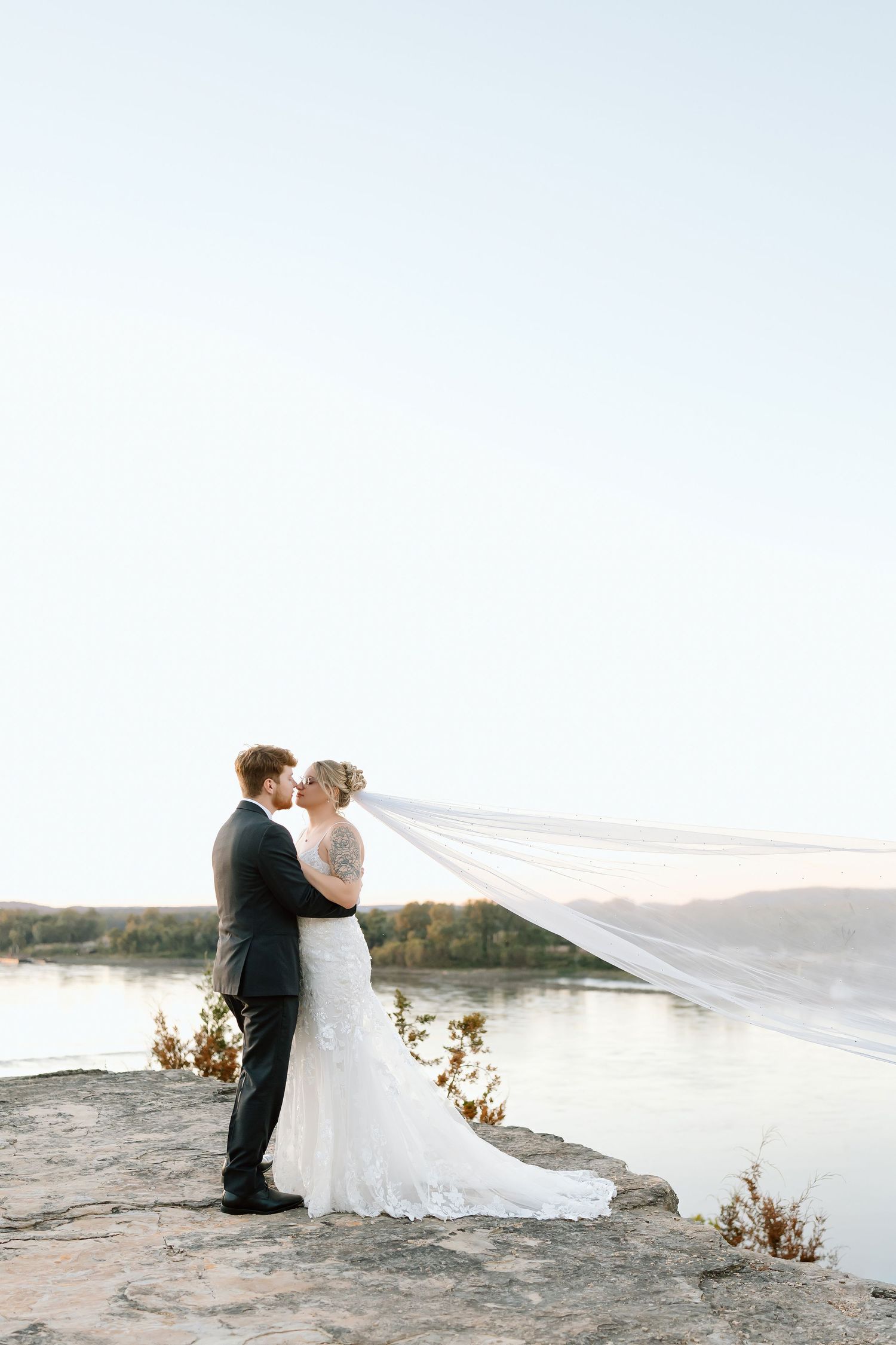 A bride and groom share an intimate moment on a scenic cliff overlooking water during sunset.