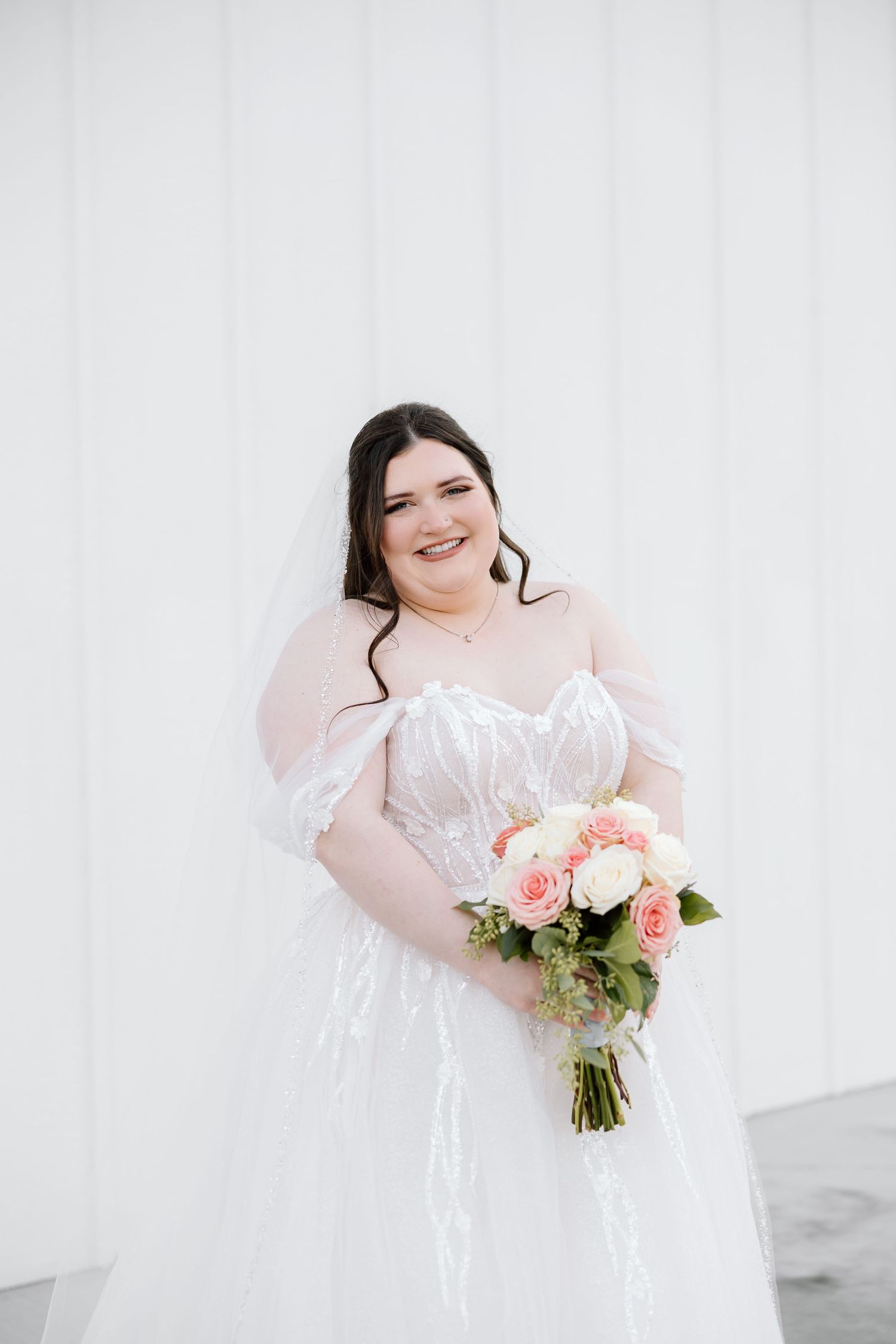 A bride in a white wedding dress holds a pink and white floral bouquet against a light background.