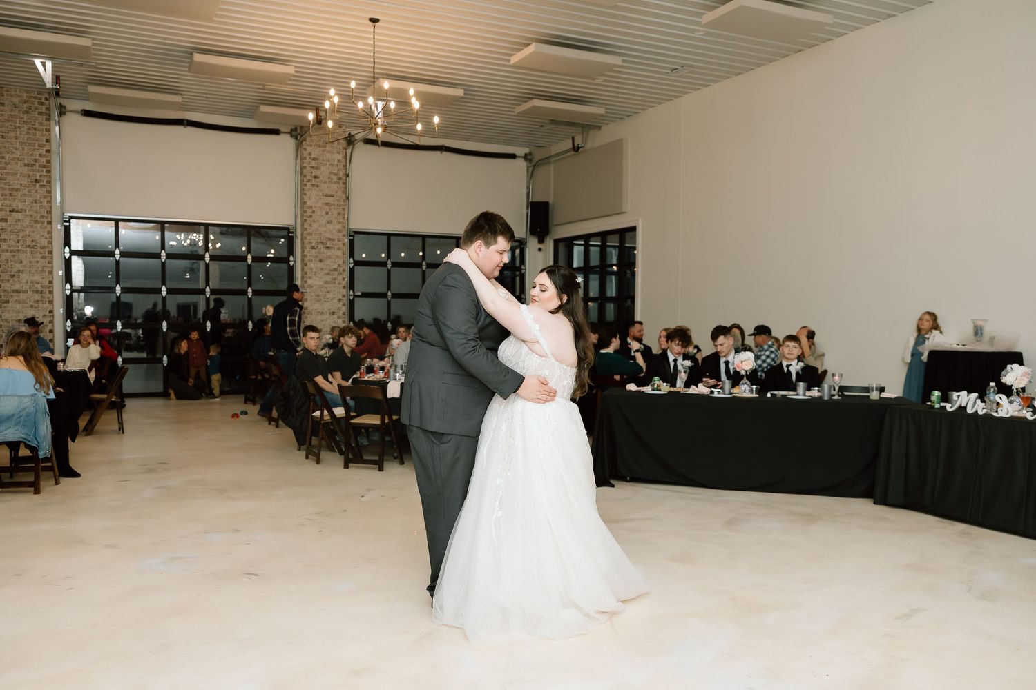 A couple shares their first dance at a wedding reception in an elegant venue with guests seated at tables in the background.