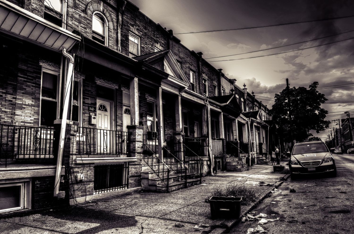 A black and white urban street scene shows a row of old brick townhouses with front porches and parked cars on a moody day.