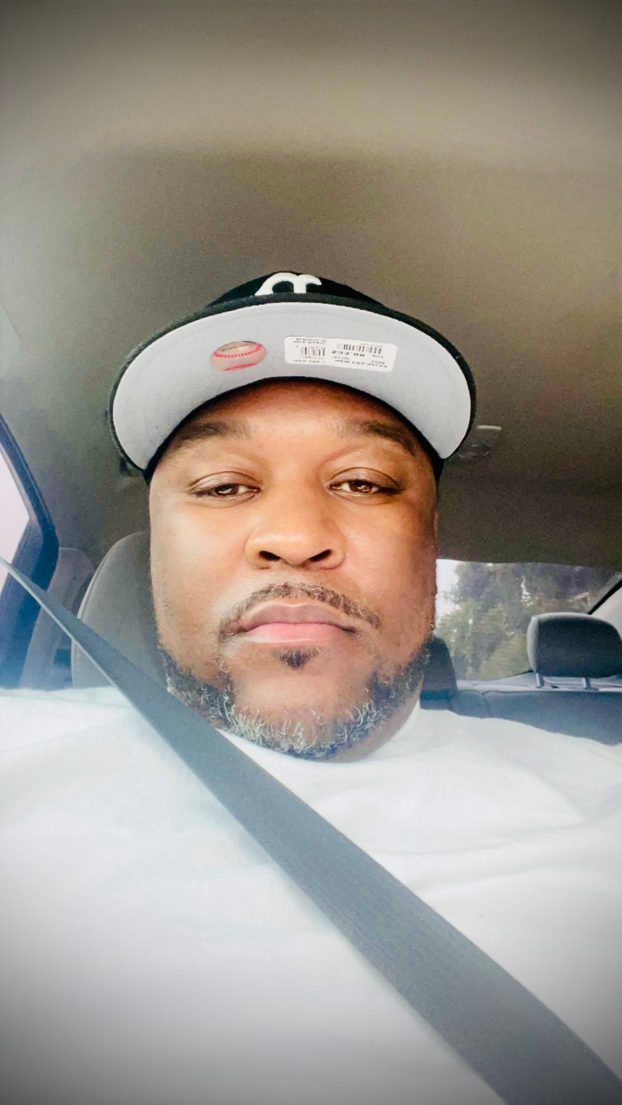 A person wearing a white and black baseball cap takes a selfie while sitting in a car.