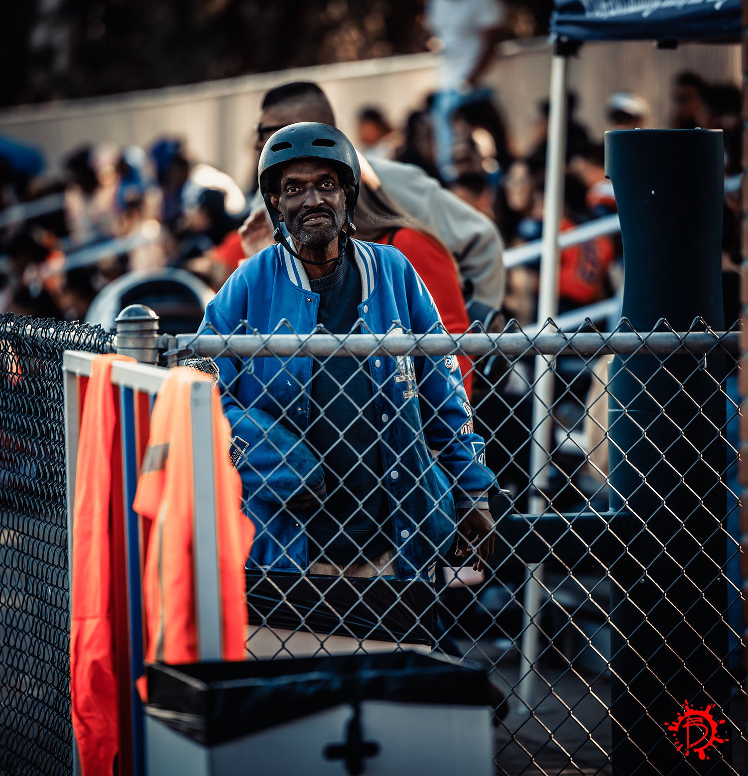 Spectator in blue jacket and cap smiles while leaning against chain link fence at outdoor sporting event.