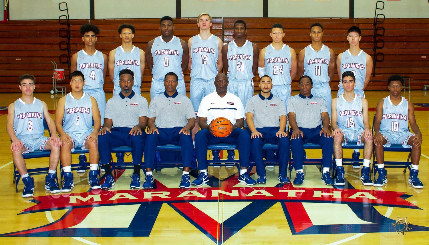 Basketball team in light blue jerseys and coaches pose for official team photo on gymnasium floor with team logo.
