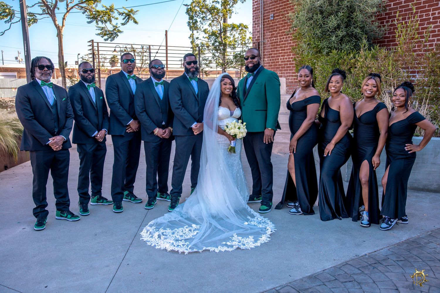 Wedding party in formal attire with groomsmen in navy suits and bridesmaids in black dresses pose outdoors by brick wall.
