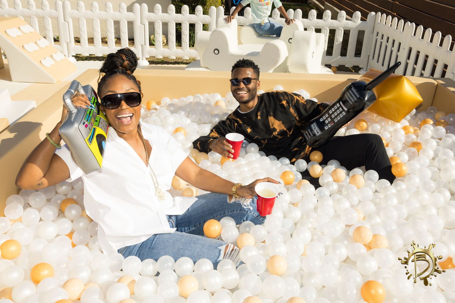 Two people relaxing in a ball pit filled with white and orange plastic balls behind a white picket fence.