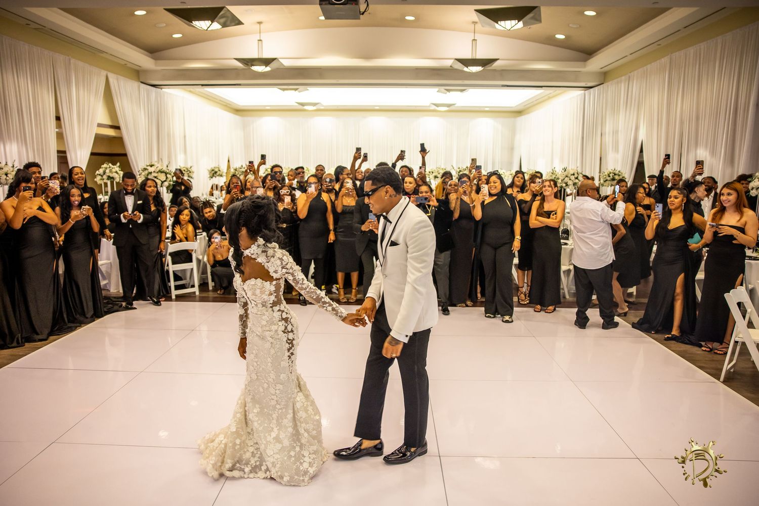 Two people share their first dance as newlyweds on a white dance floor surrounded by wedding guests in formal attire.