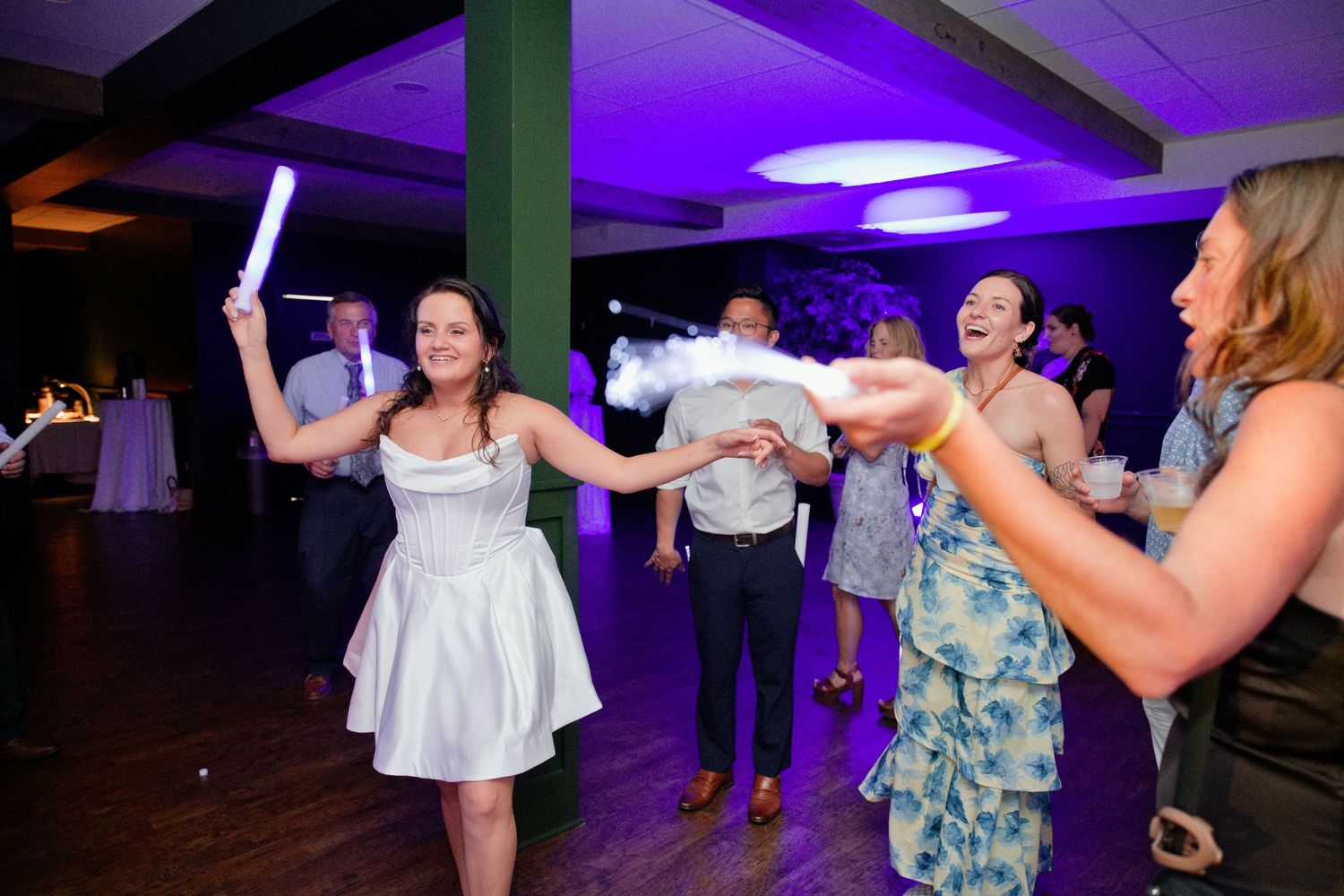 Bride in white dress dancing with guests at wedding reception under purple lights holding glow stick.
