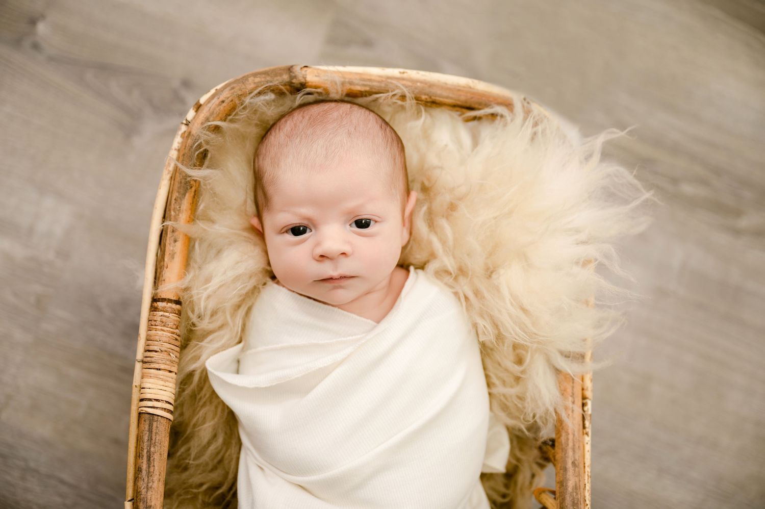 A small infant swaddled in white fabric rests in a wicker chair with soft creamy fur padding.