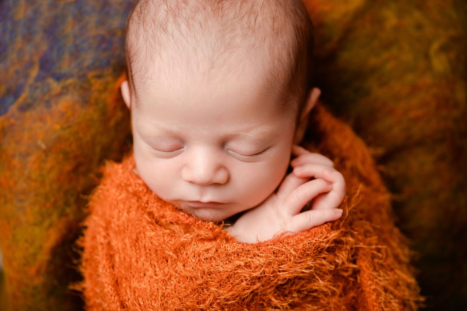 Sleeping newborn wrapped in soft orange blanket on brown textured backdrop and nestled in cozy position.