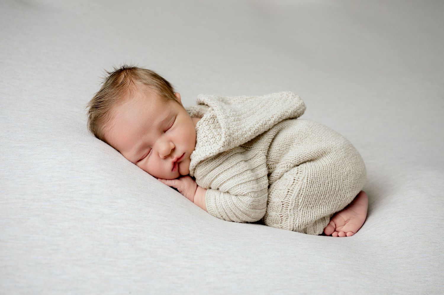 Minnesota Best newborn photographer - A sleeping newborn baby dressed in a cream knit sweater lies peacefully on soft white fabric.
