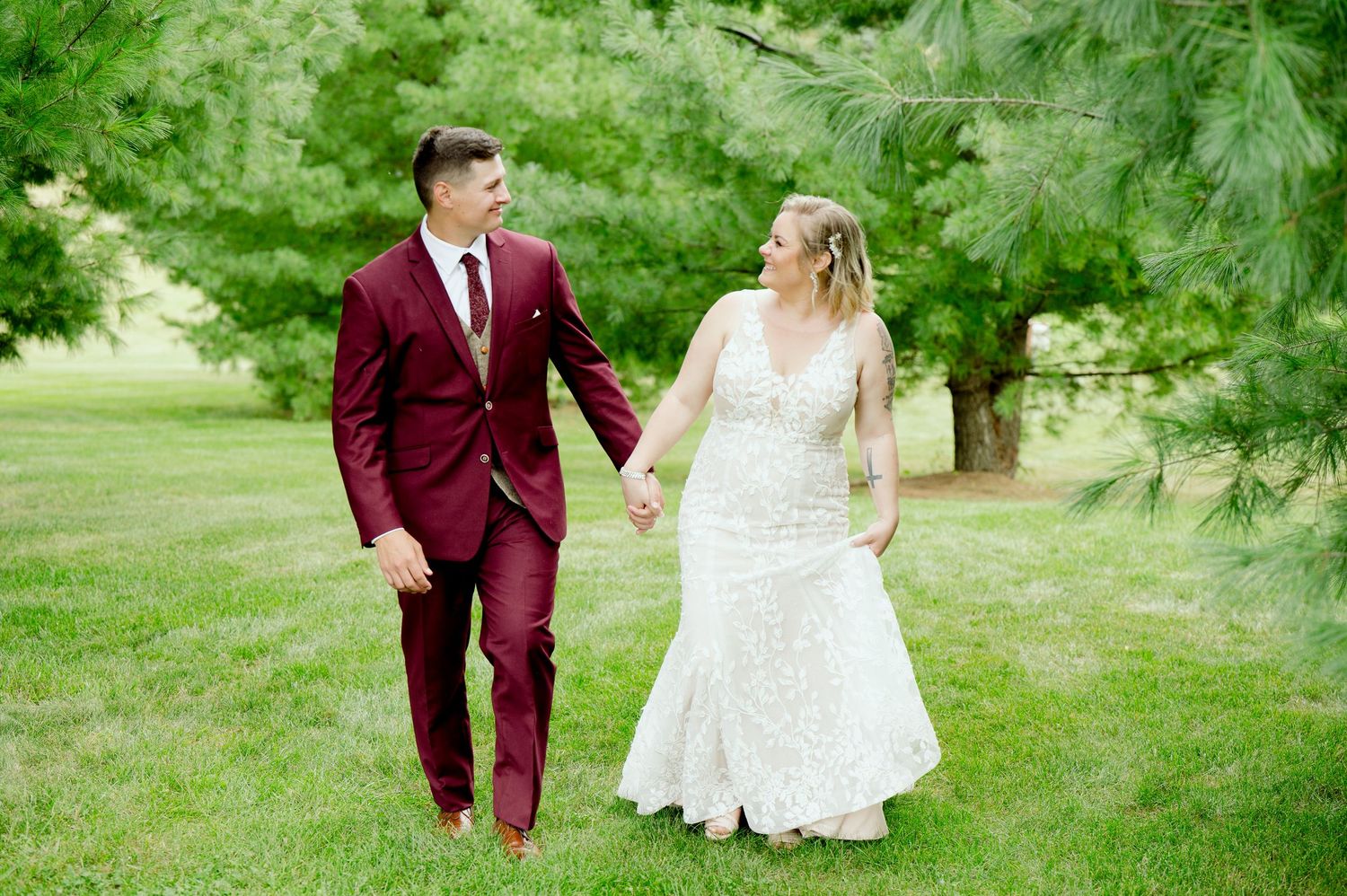 Couple walks hand in hand across lawn, bride in lace dress and groom in burgundy suit.