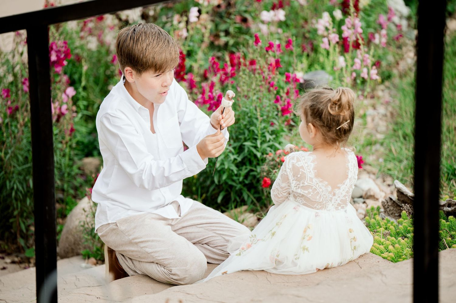 Children sitting on outdoor steps surrounded by pink flowers in a garden setting.
