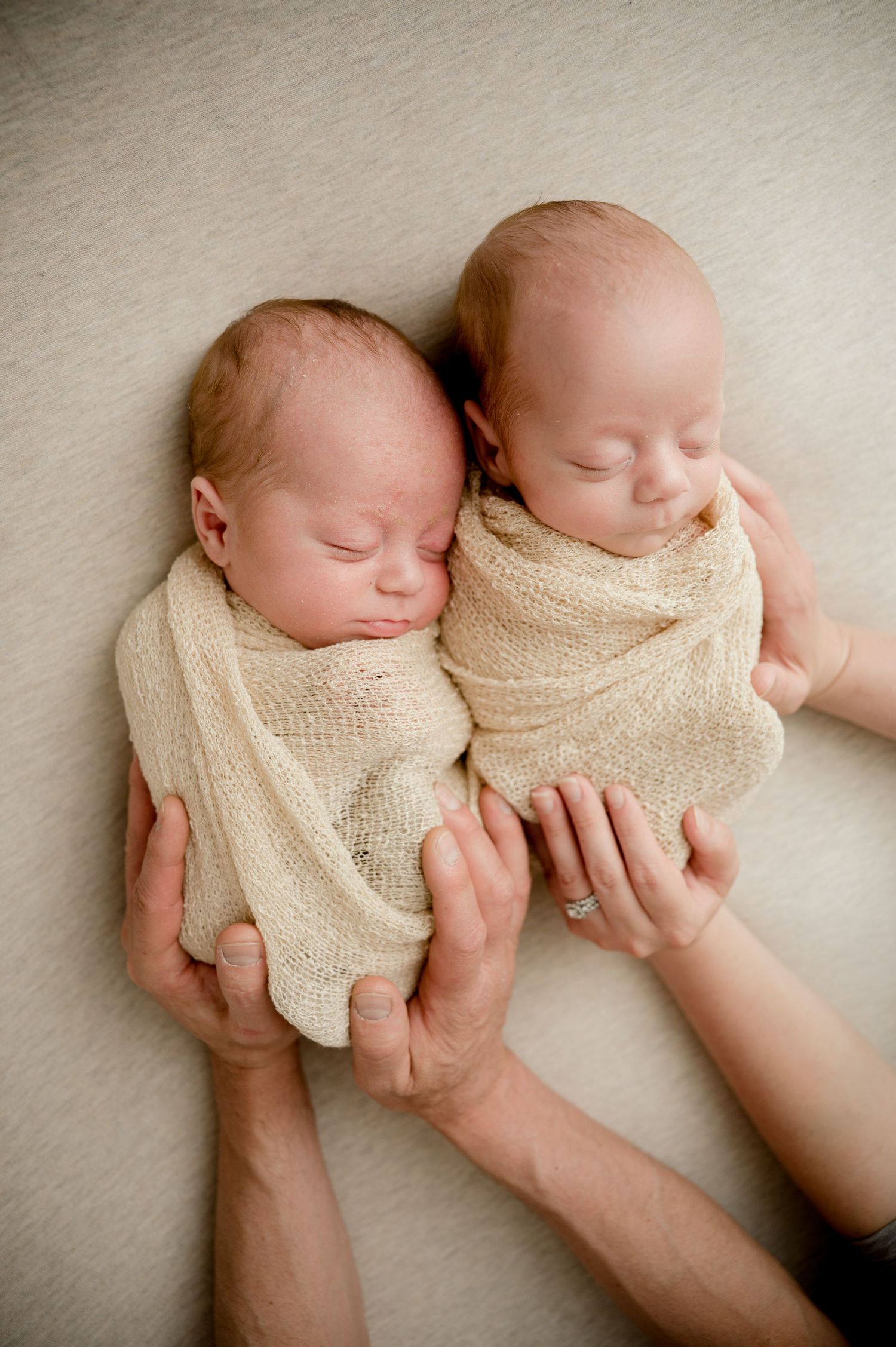 Two swaddled newborn twins sleeping side by side, gently cradled in adult hands on a soft neutral background.