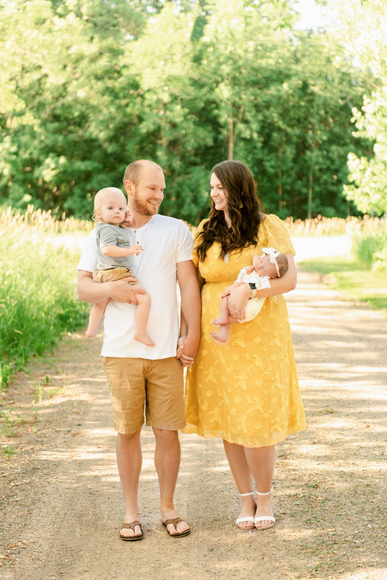 St Cloud newborn Photos of Young family poses outdoors on a sunny dirt path, parents holding a toddler and newborn baby.