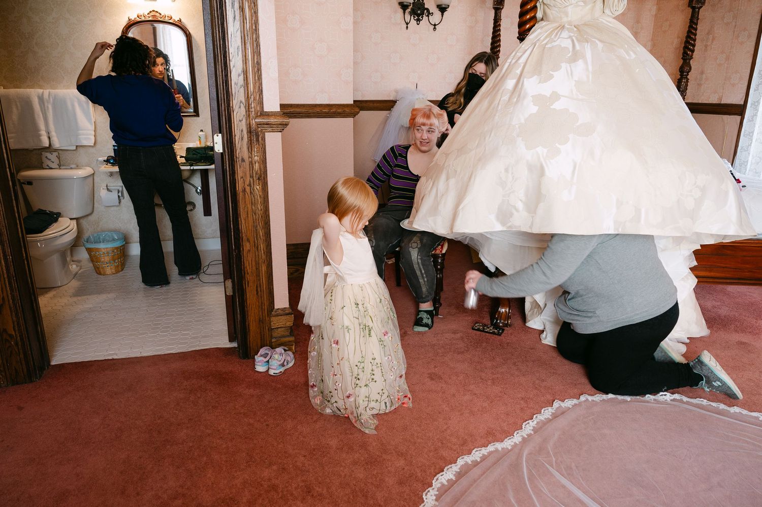 Central MN Wedding Photos | A flower girl watches as family members help adjust a bride's white wedding gown in a carpeted dressing room.