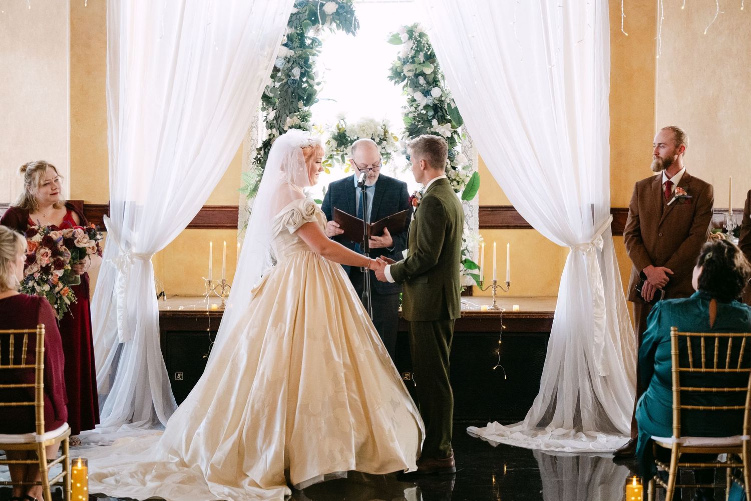 Central MN Wedding Photos | Bride and groom exchanging vows at elegant indoor ceremony with white drapes and floral arch.
