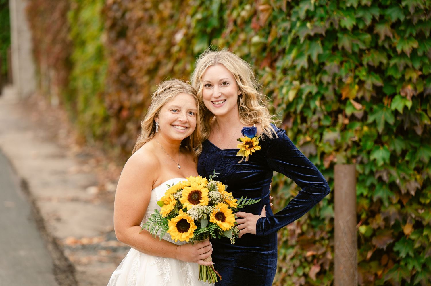Central MN Wedding Photos | Bride and guest pose together with sunflower bouquet in front of colorful ivy wall.