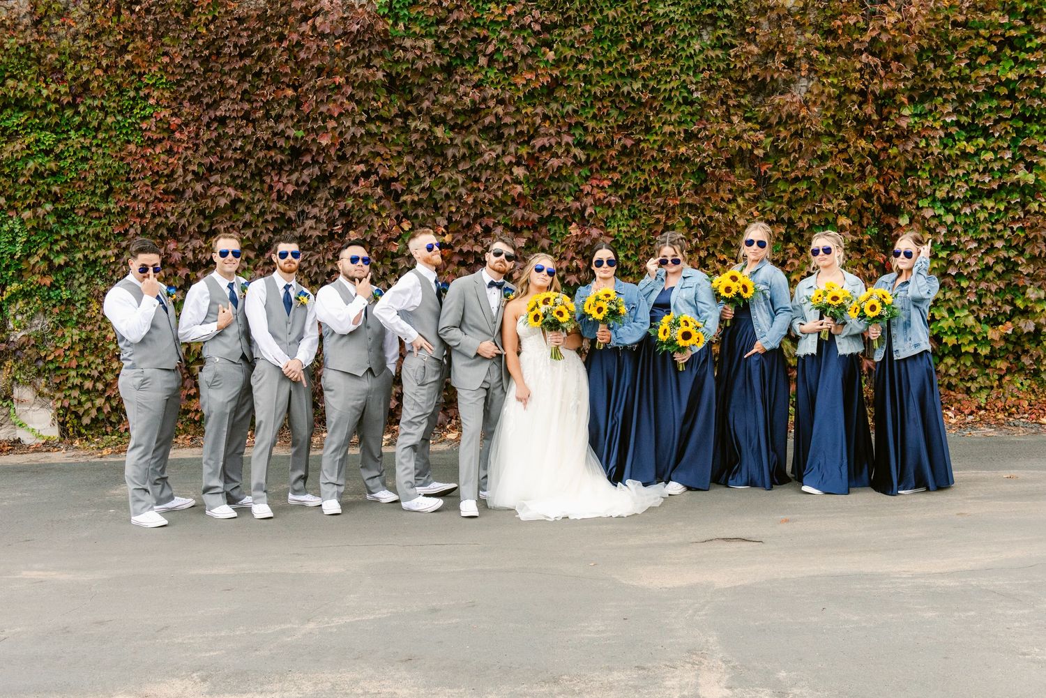 Central MN Wedding Photos | Wedding party in gray suits and navy dresses wearing sunglasses holding sunflowers against ivy wall.