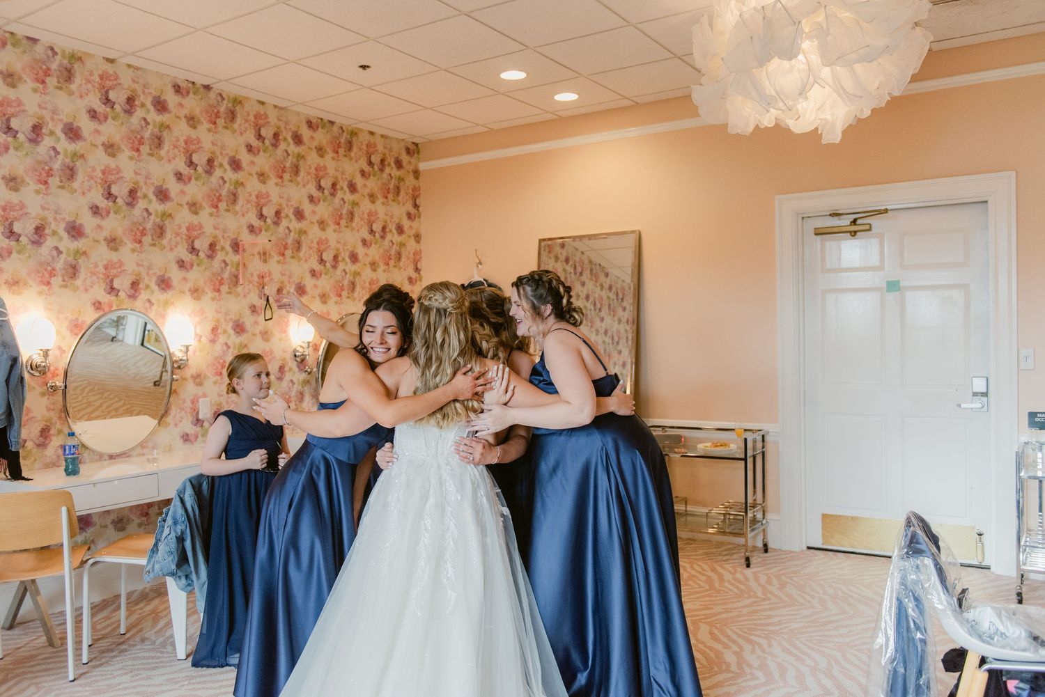 Central MN Wedding Photos | Bride and bridesmaids in navy blue dresses share a group hug in a floral wallpapered bridal suite.