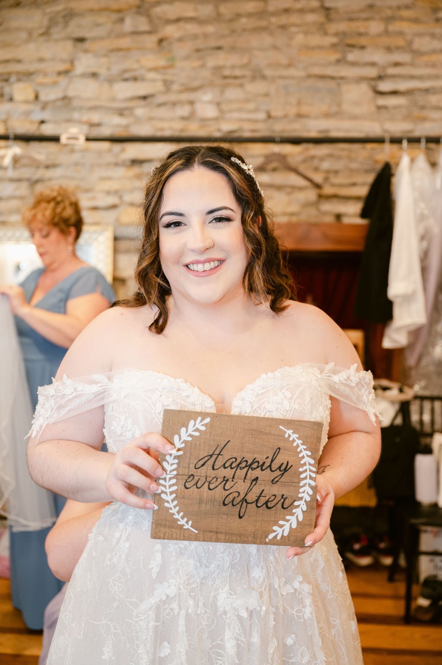 Central MN Wedding Photos - Smiling bride in off-shoulder lace gown holds a rustic Happily Ever After sign at her bridal fitting.