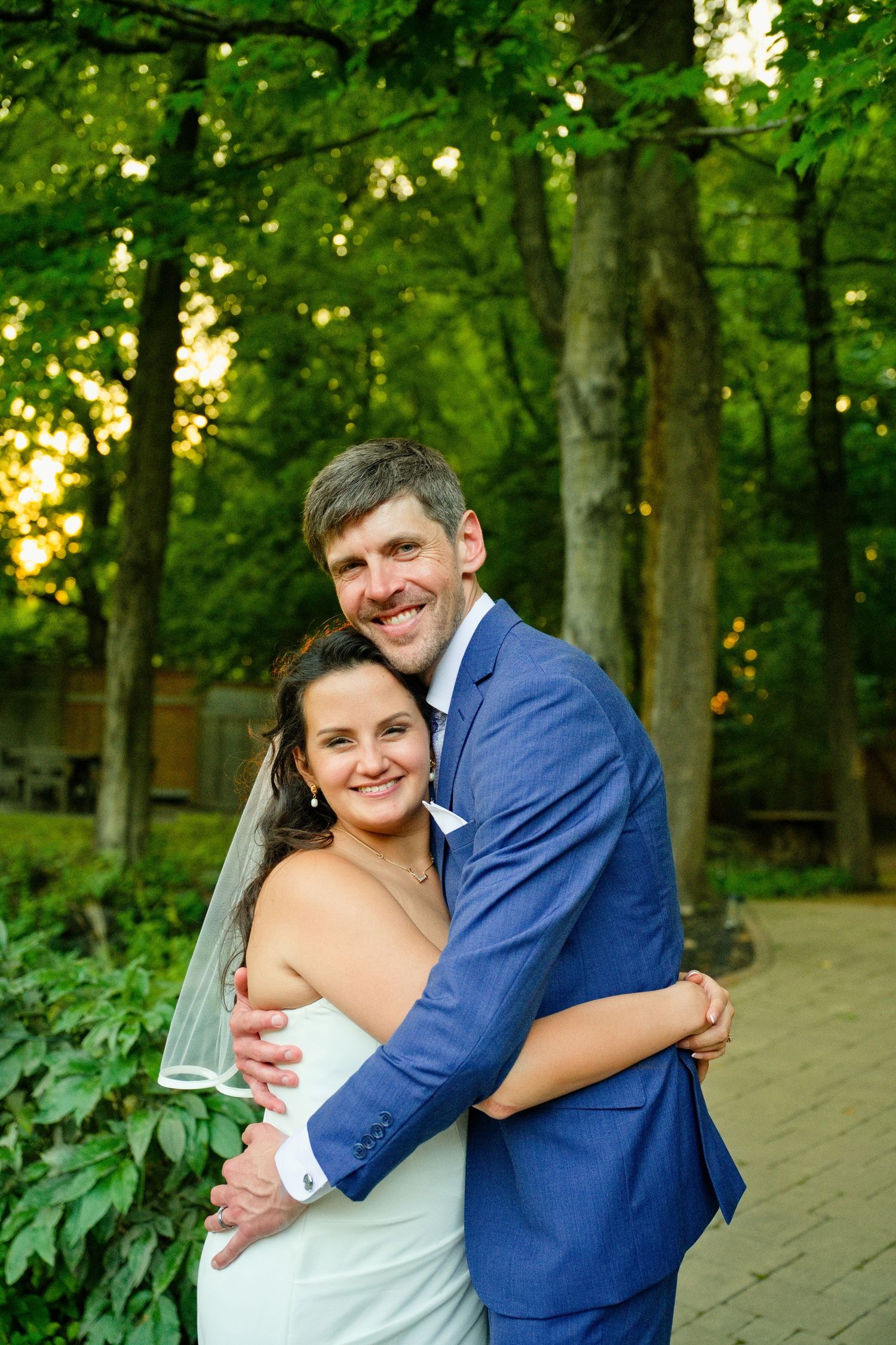 A smiling bride and groom embrace outdoors in a lush green park setting, she in white gown with veil, he in blue suit.