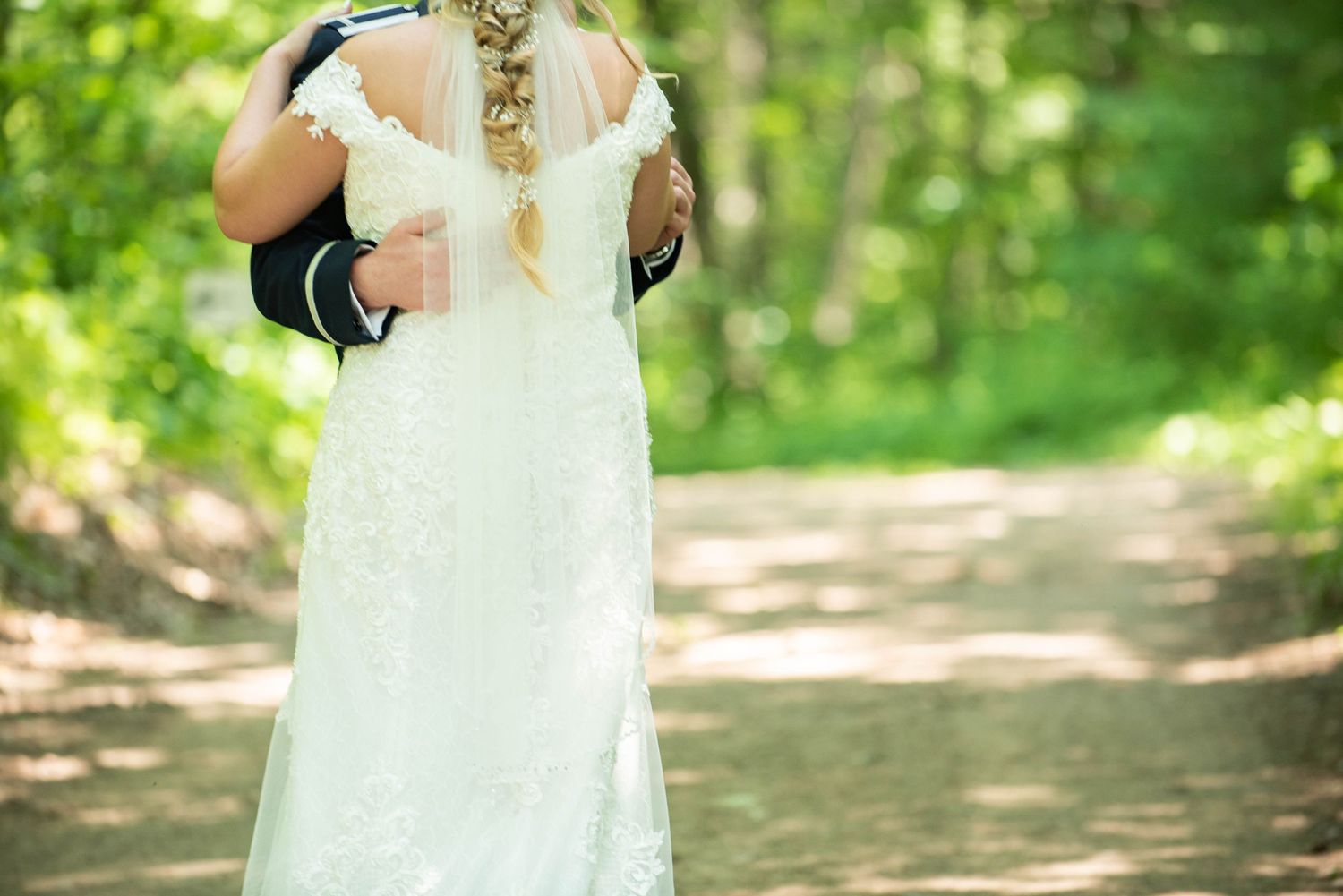 Couple in formal wedding attire holding hands while walking through a sunlit forest path. Zimmerman MN Weddings