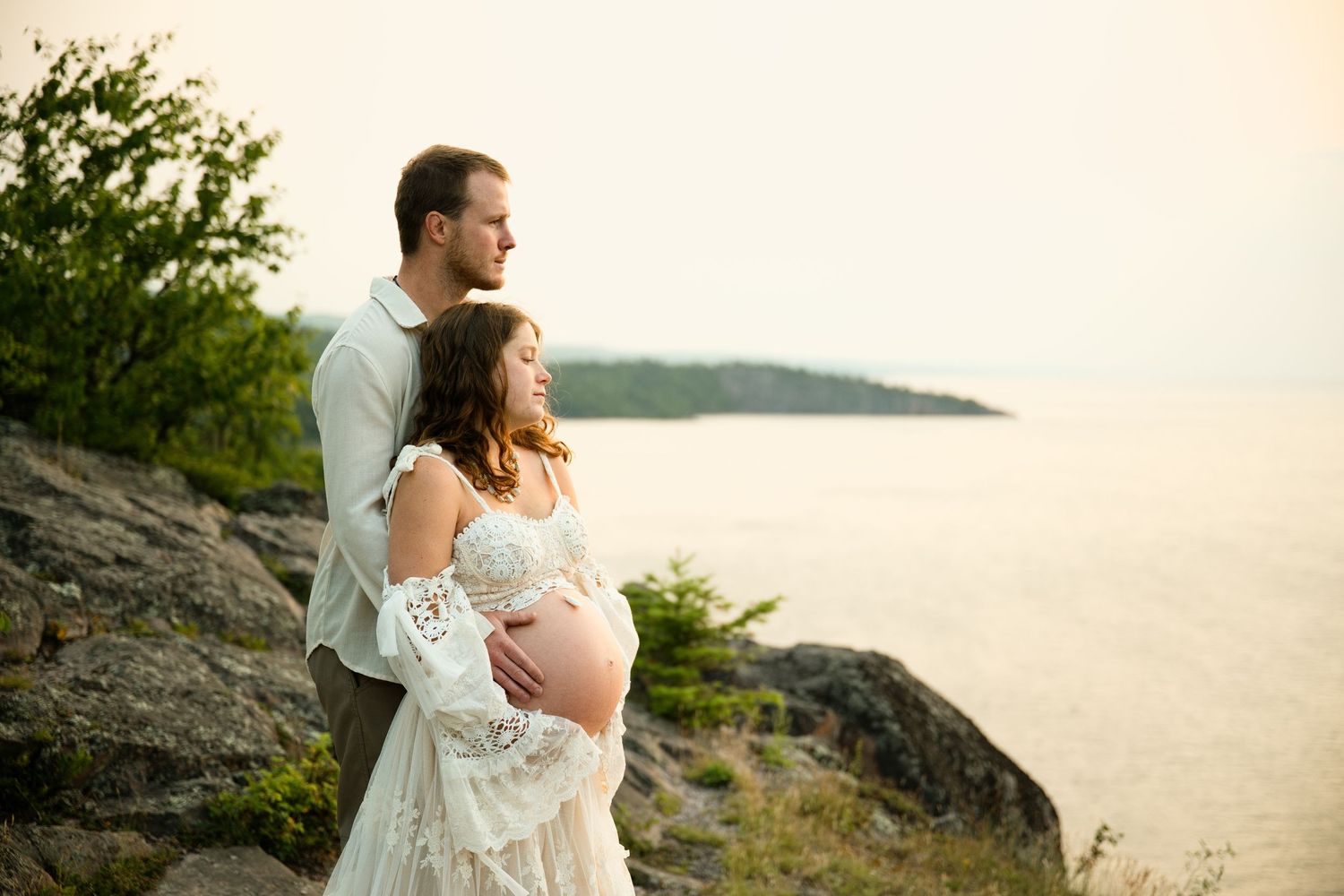 Pregnant couple standing on rocky cliffside overlooking calm water at sunset, woman in lace maternity gown.