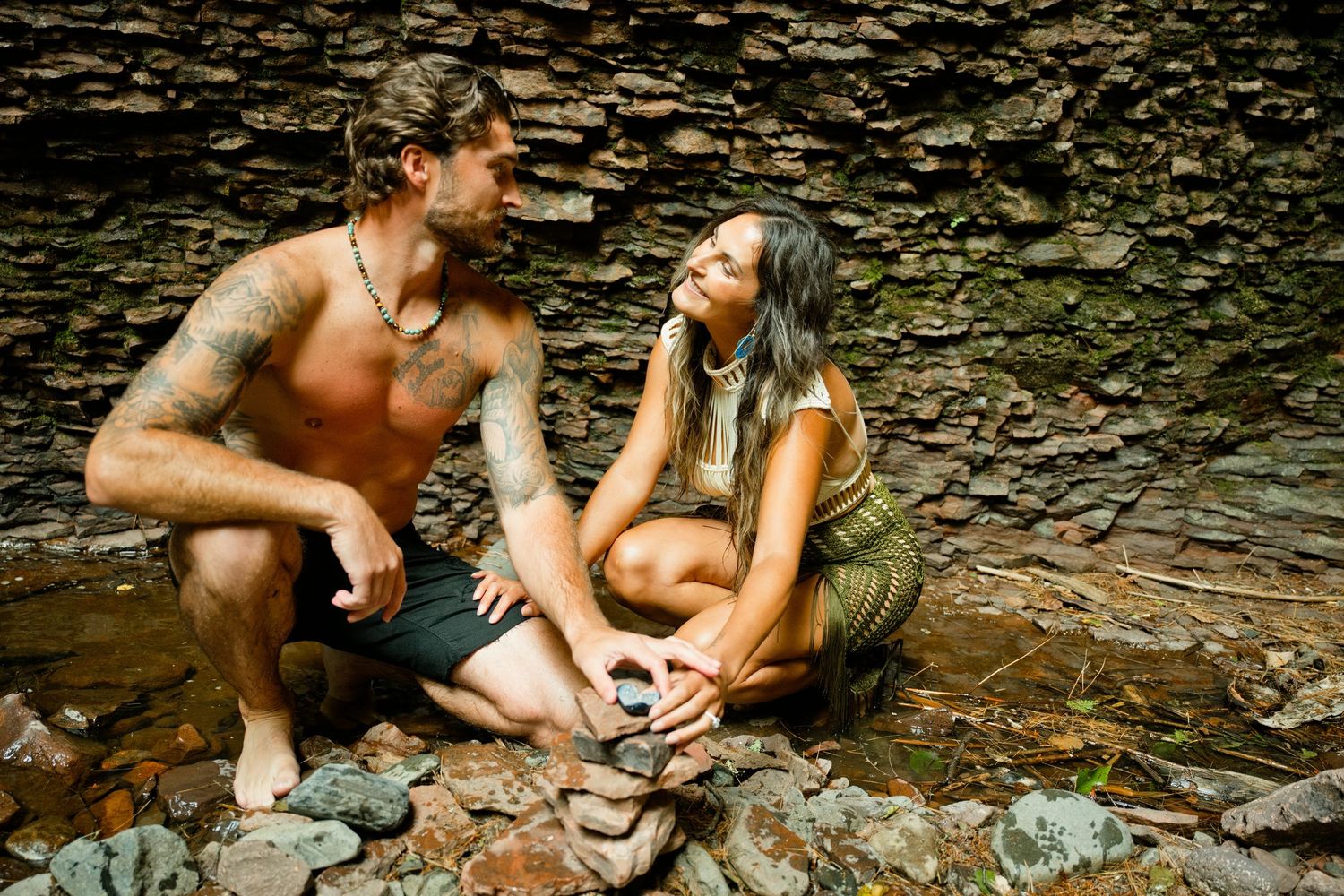 Shirtless tattooed man and woman in olive dress sit together on rocks near cave wall.