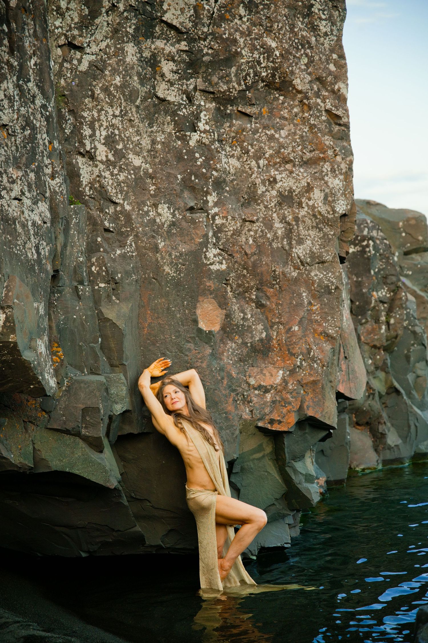 Woman in sheer dress poses gracefully against rugged rock face by water.