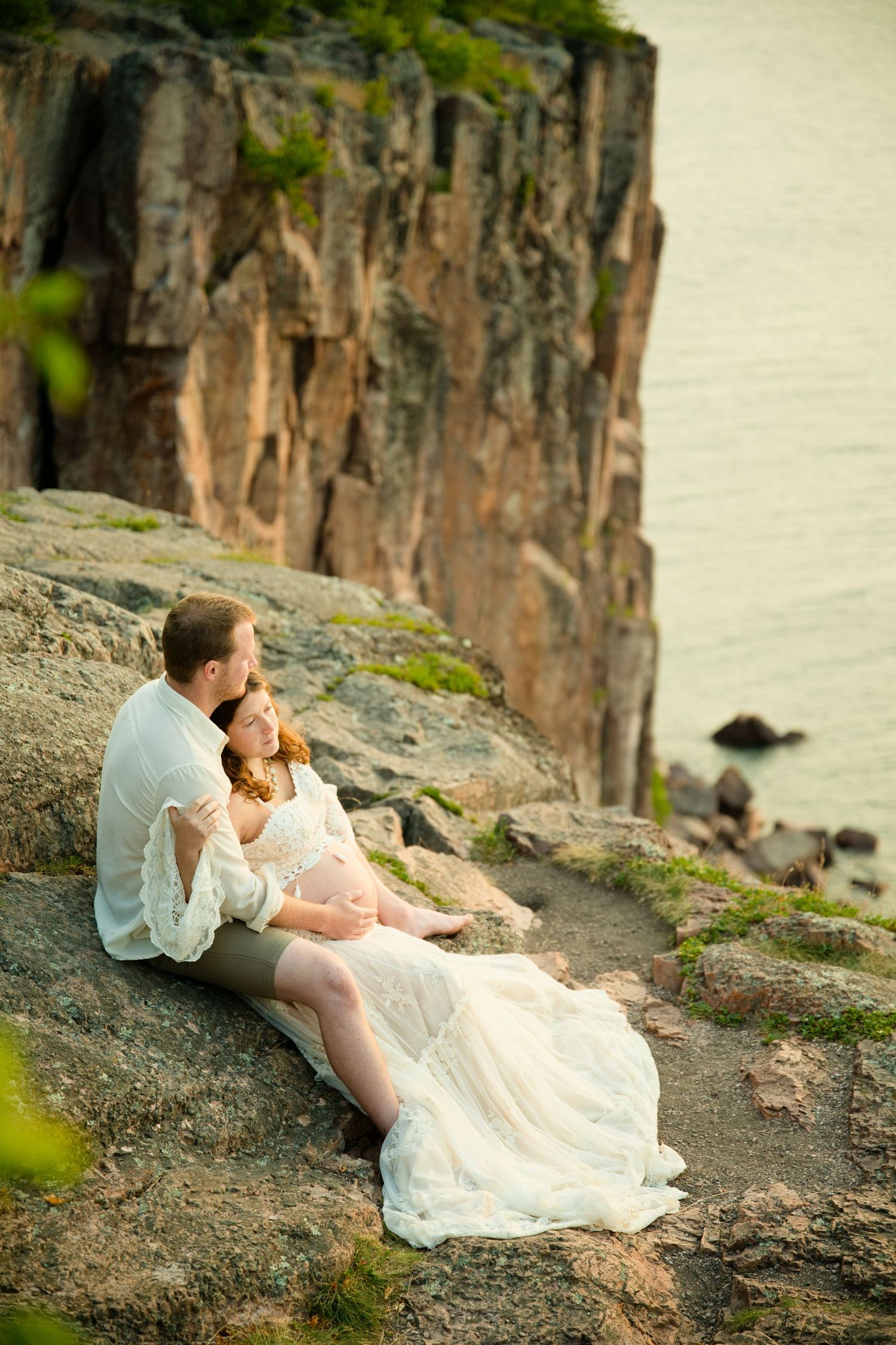 Couple sitting on rocky cliff edge by water, woman in white lace dress, man in white shirt, sharing a romantic moment.