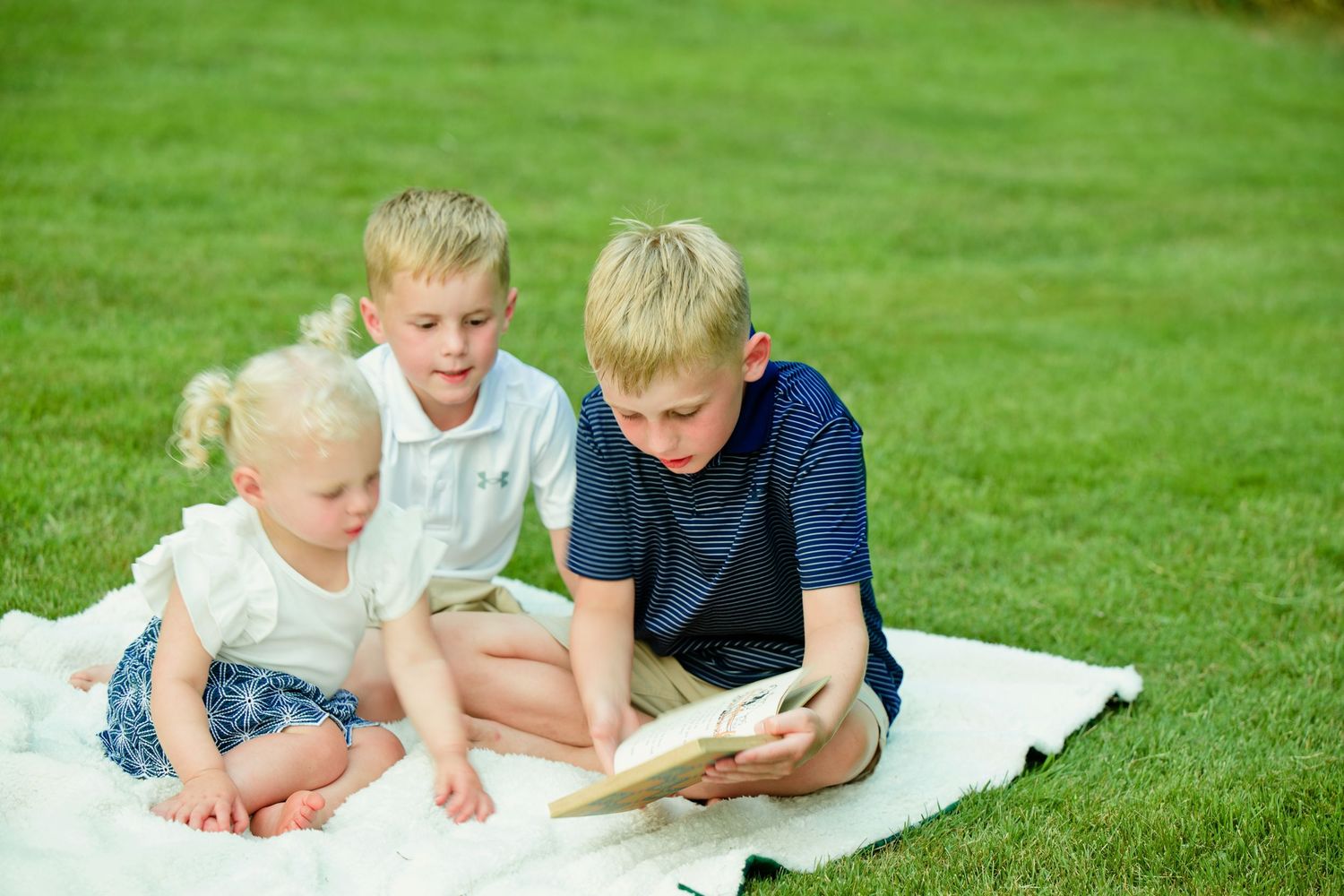 Three young blonde children reading a book together on a white blanket in a lush green grass yard.