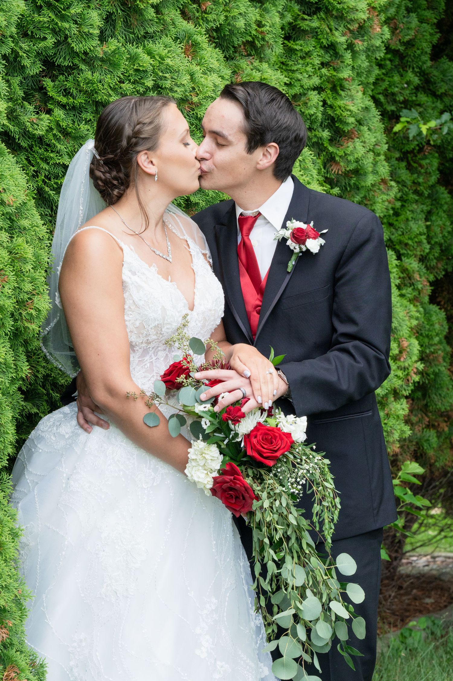 Newlyweds share romantic kiss while bride holds cascading red rose bouquet in front of lush green hedges.