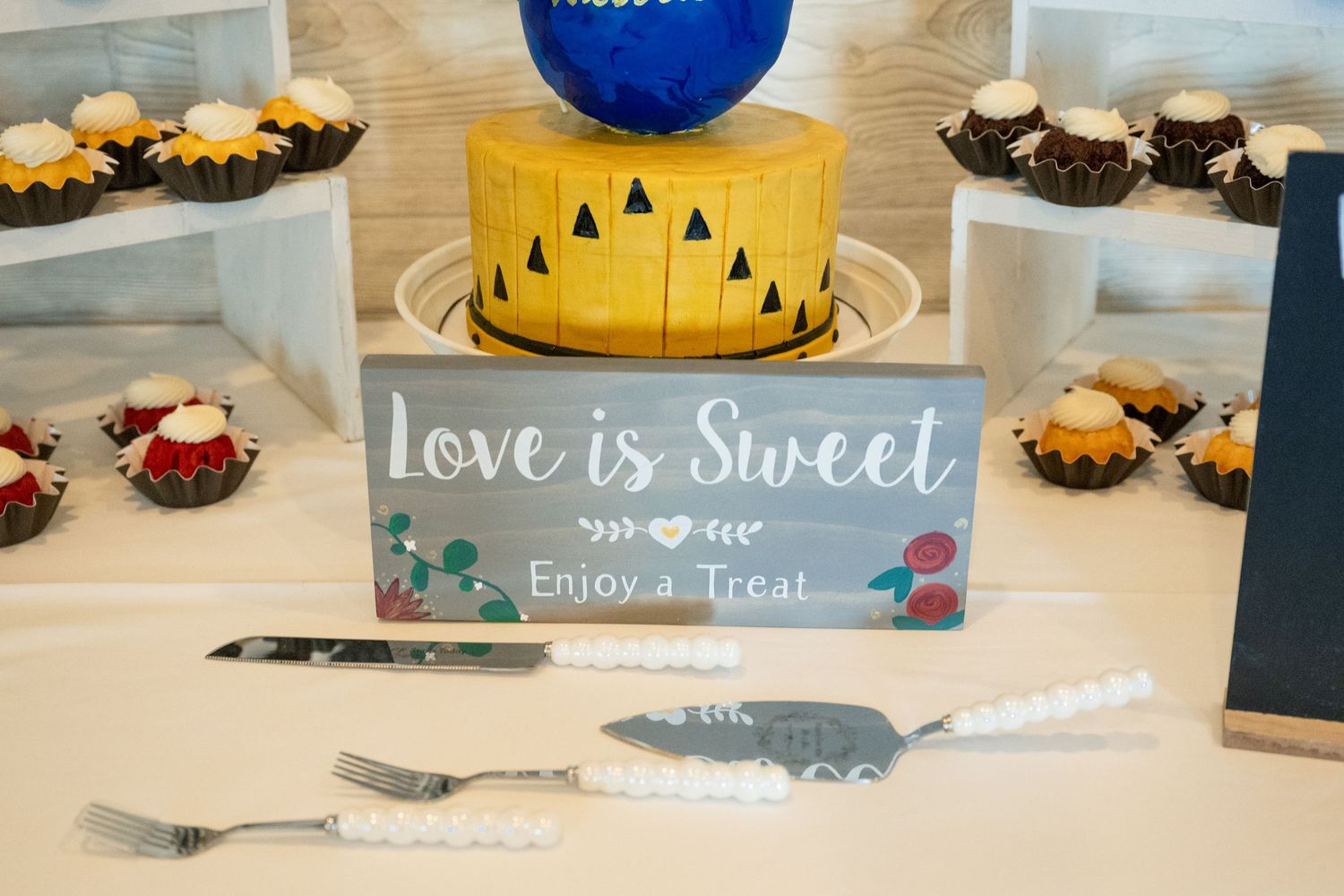 Wedding dessert table featuring a yellow cake, cupcakes, and a blue Love is Sweet enjoy a treat sign.