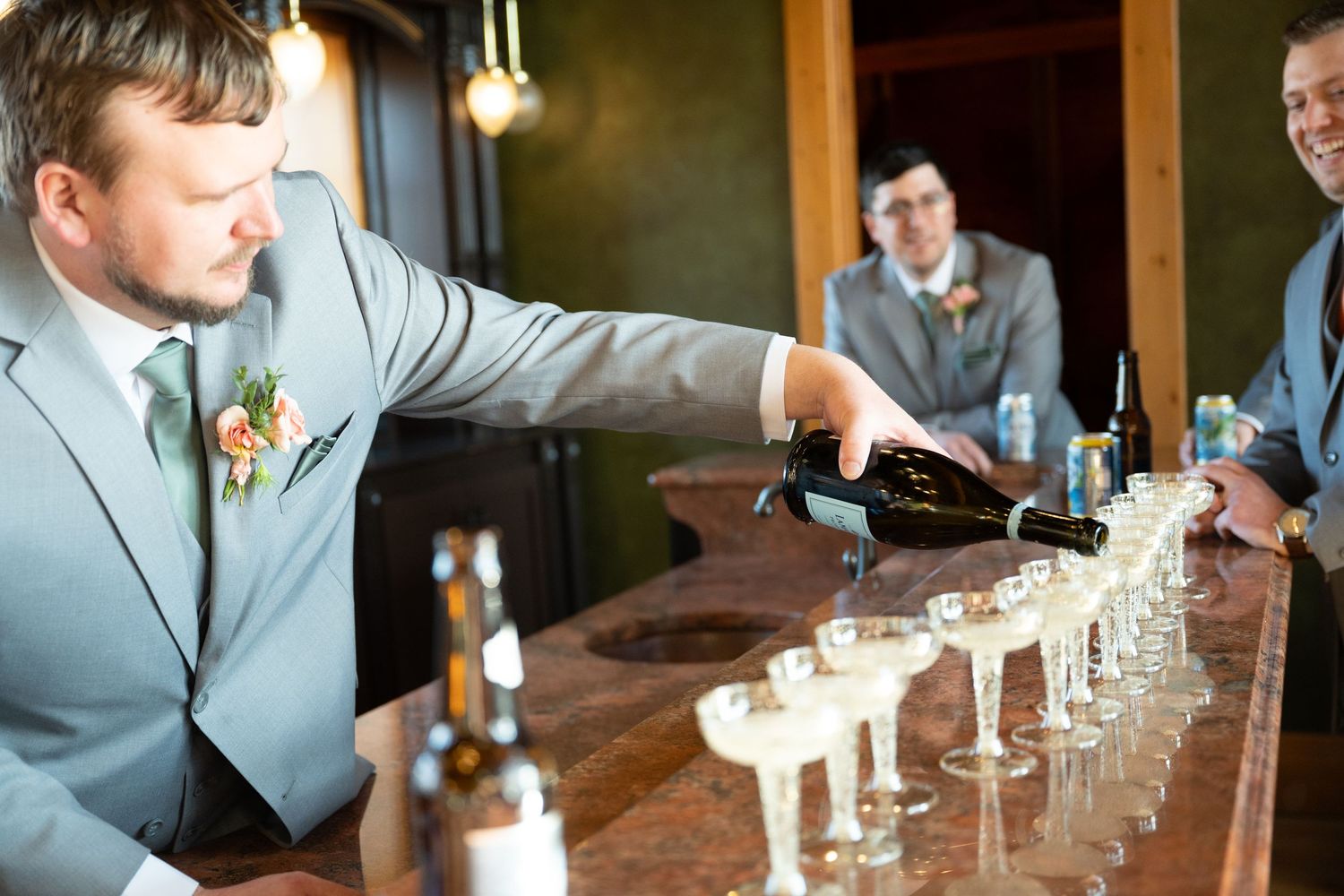 Groomsman in gray suit pours champagne into a row of glasses at a wedding bar reception.