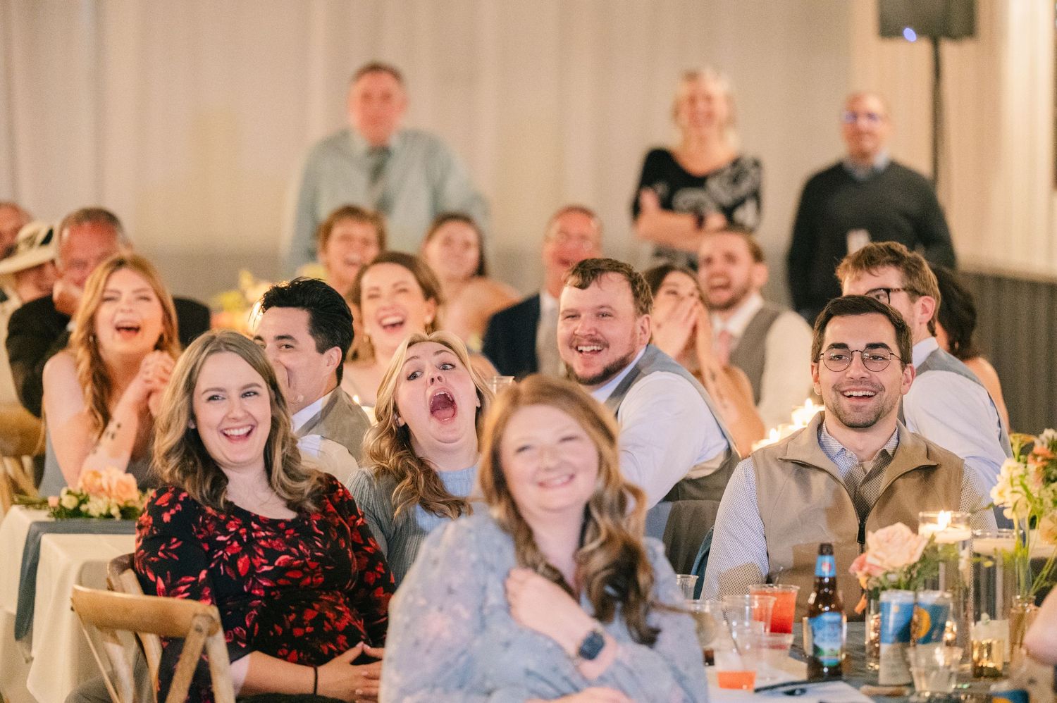 Joyful wedding guests laugh and react during reception speeches at elegantly decorated reception hall.