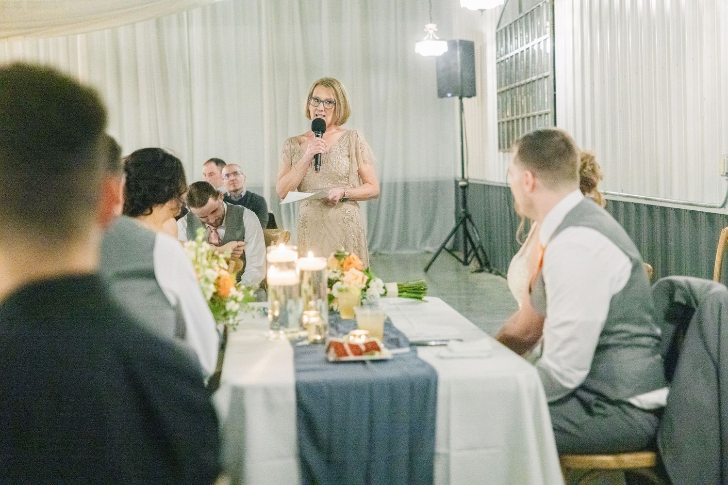 A woman in a gold dress gives a speech at a wedding reception with guests listening.