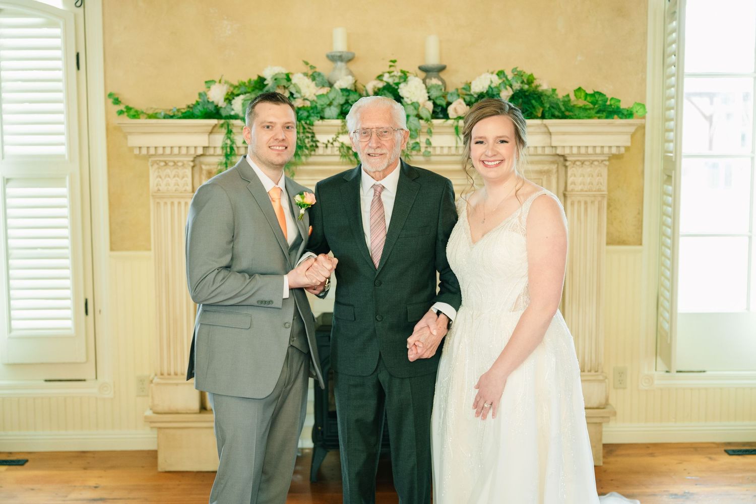 Bride and groom pose with elderly officiant before floral fireplace mantel at wedding ceremony.