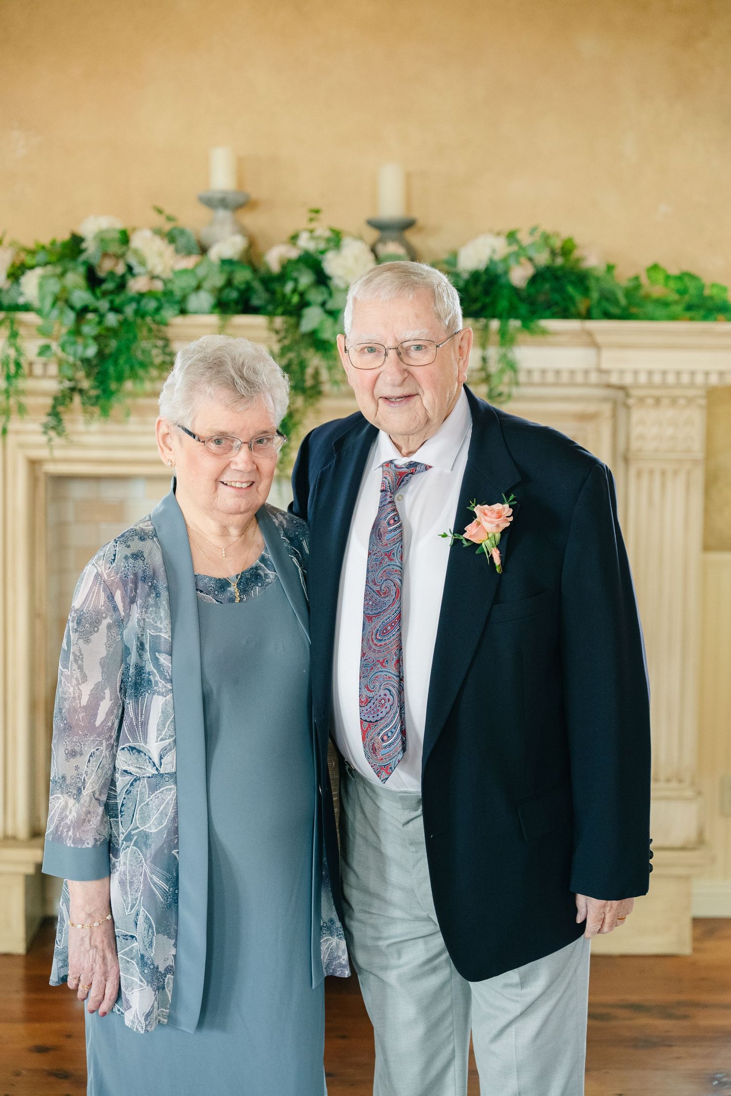 Elderly couple posing at wedding, man wearing boutonniere and navy blazer with tie.