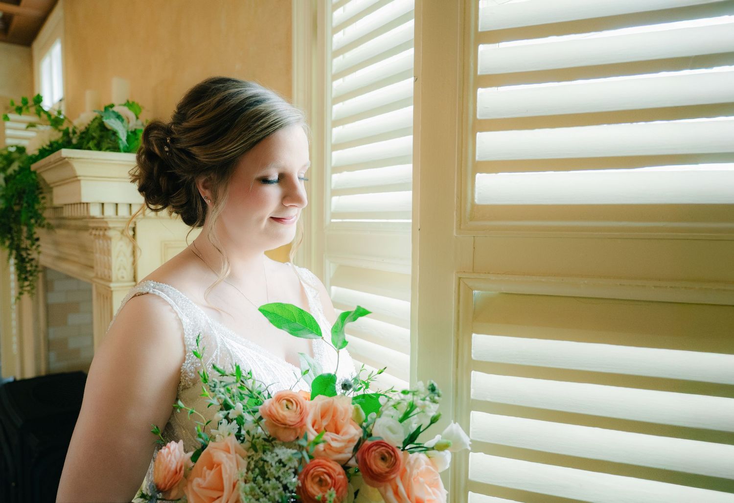 Bride in lace wedding gown holding peach rose bouquet while gazing out window by fireplace.