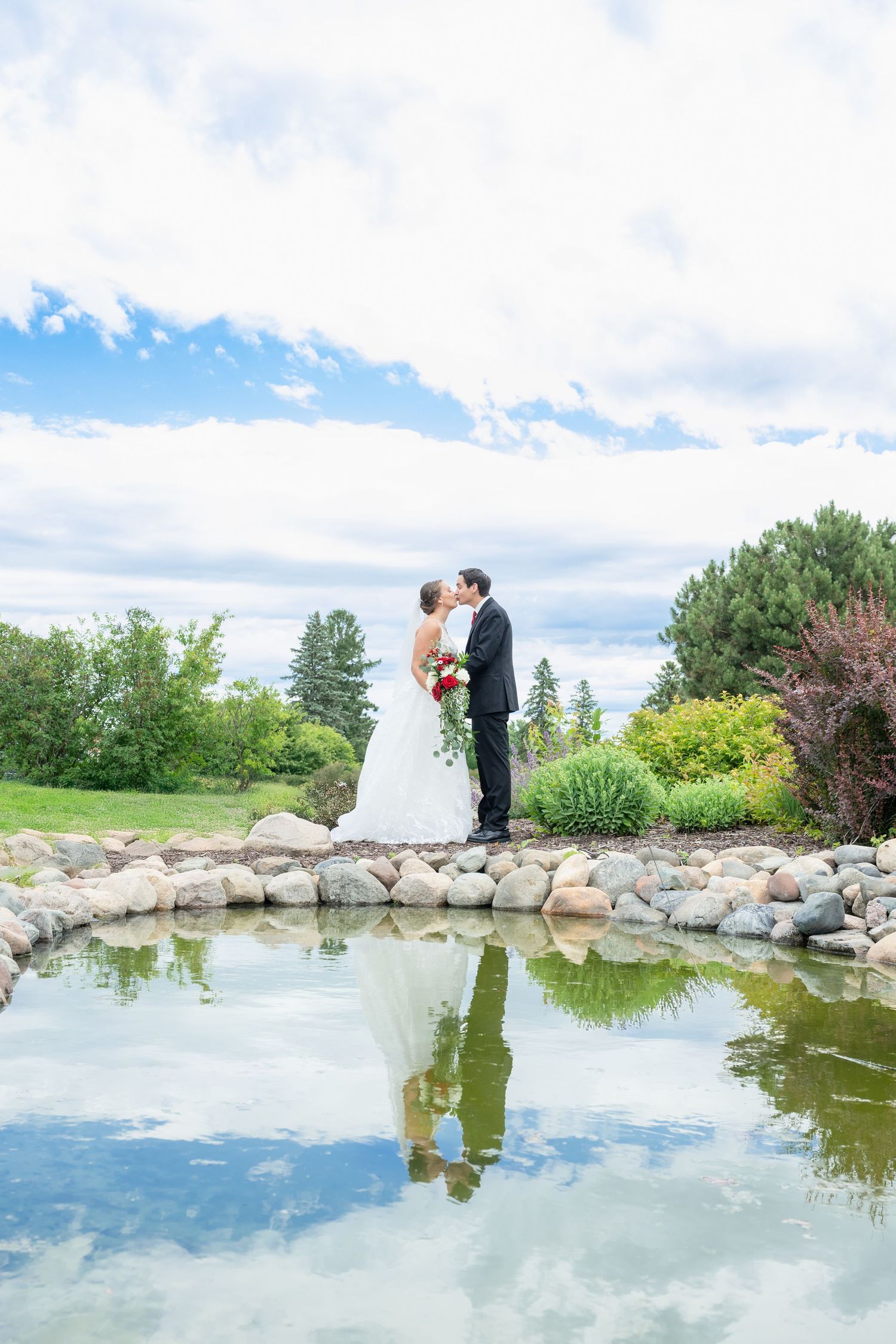 Bride and groom share a kiss beside a reflective pond in a lush garden on their wedding day.